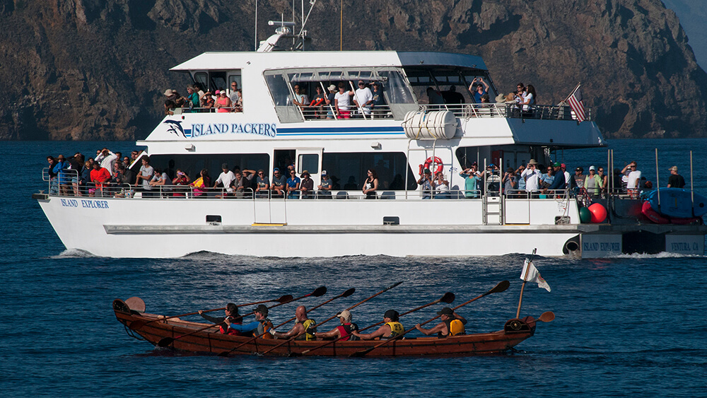 People on a tomol paddle next to a large power vessel